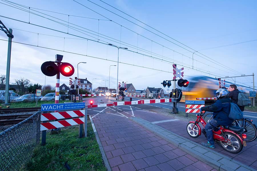 Bewaakte spoorwegovergang met slagboom, bel en knipperlicht. Een brommer en fietser wachten tot de trein voorbij is.