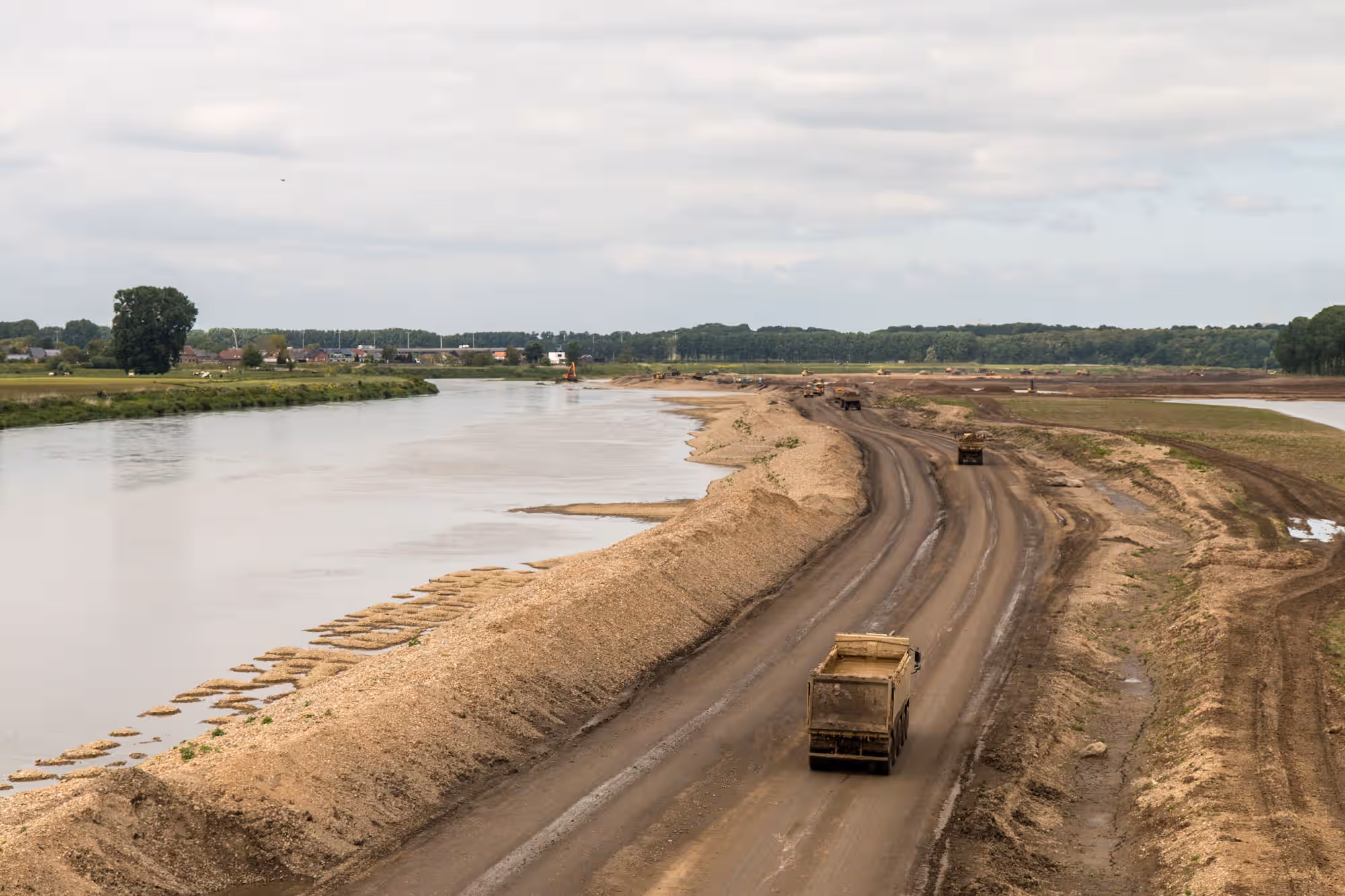 Een bouwweg in de uiterwaarden langs een rivier met daarop grote bouwvrachtwagens